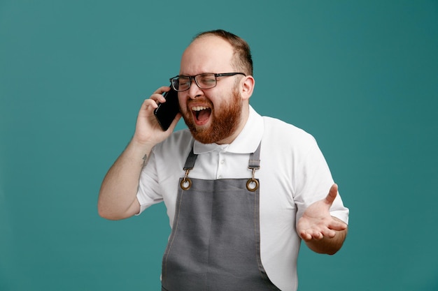 Gestresster junger Friseur in Uniform und Brille, der am Telefon schreit und mit geschlossenen Augen schreit und leere Hand isoliert auf blauem Hintergrund zeigt