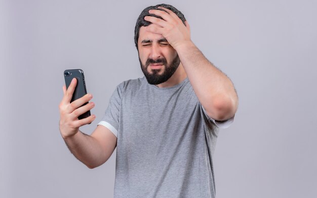 Gestresster junger gutaussehender Mann, der Handy hält Hand auf Kopf mit geschlossenen Augen lokalisiert auf weißer Wand hält