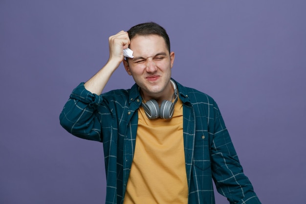 Gestresster junger männlicher Student, der Kopfhörer um den Hals trägt und den Kopf mit zerrissenem und zerdrücktem Prüfungspapier berührt, mit geschlossenen Augen, isoliert auf violettem Hintergrund