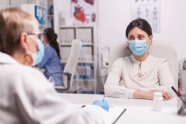 Gestresster junger Patient mit Gesichtsmaske gegen Covid-19 während der Konsultation mit dem Oberarzt im Krankenhausbüro. Erschrockene Frau.