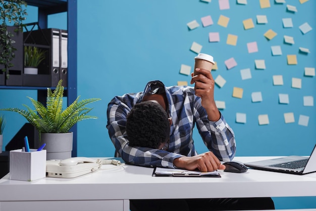 Gestresster und ermüdeter Teamleiter mit Burnout-Syndrom, der in einem modernen Büroarbeitsplatz sitzt. Müder und erschöpfter Büroangestellter der Agentur, der wegen Überstunden am Schreibtisch einschläft