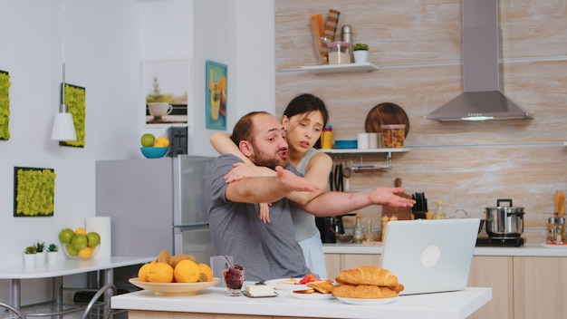 Gestresster Unternehmer, der an einem Laptop arbeitet, der auf dem Küchentisch sitzt, während seine Frau das Frühstück kocht. Unglücklicher, gestresster, frustrierter wütender negativer und verärgerter Freiberufler im Pyjama, der morgens schreit