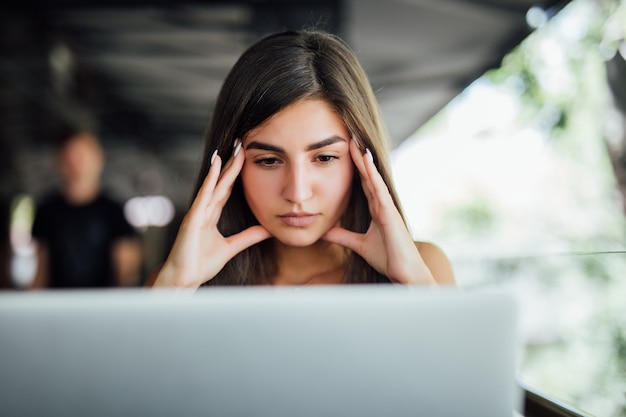 Gestresstes beschäftigtes müdes junges Mädchen bei der Arbeit an ihrem Bericht vor dem Laptop im Sommercafé