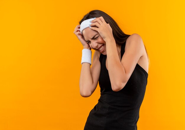 Gestresstes junges hübsches sportliches Mädchen, das Stirnband und Armband trägt, die Hände auf Kopf mit geschlossenen Augen lokalisiert auf orange Raum setzen