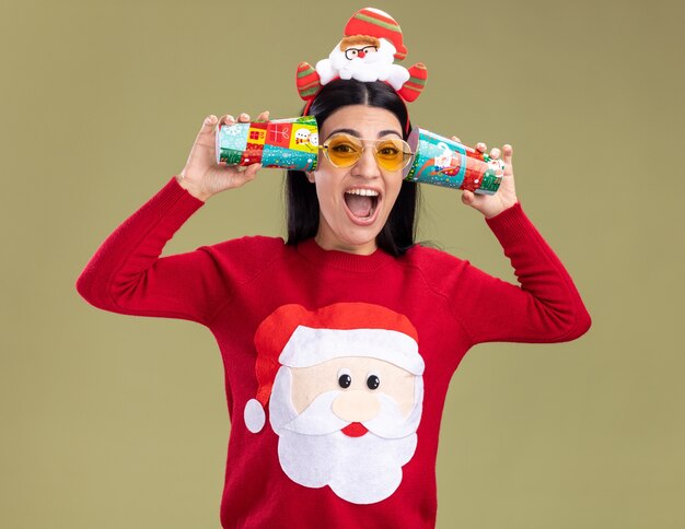 Kostenloses Foto gestresstes junges kaukasisches mädchen mit weihnachtsmann-stirnband und pullover mit brille, das plastikweihnachtsbecher neben den ohren hält und gesprächen zuhört, die einzeln auf olivgrüner wand schreien?