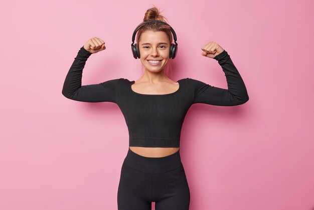 Gesunde motivierte Frau hebt die Arme und zeigt Bizeps nach regelmäßigem Training zeigt ihre sportlichen Leistungen hört Musik über Kopfhörer Posen vor rosa Hintergrund Menschen- und Kraftkonzept