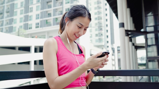 Gesunde schöne junge asiatische Athletenfrau, die Smartphone für verwendet, hören Musik beim Laufen