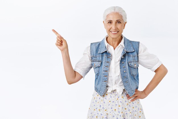 Gesunde und fröhlich lächelnde alte stilvolle Dame mit grauem Haar trägt trendiges Denim-Westenkleid, das nach links zeigt, halten Sie die Hand auf der Hüfte und grinsen Sie selbstbewusst, zeigen Sie die beste Wahl, geben Sie Ratschläge, empfehlen Sie das Produkt