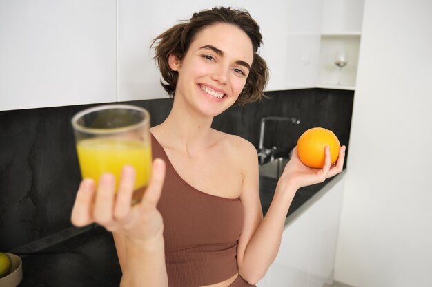 Gesunder Körper und Wohlbefinden lächelndes Fitness-Mädchen, das Ihnen ein Glas frischen Saft mit einer Orange in der Hand gibt