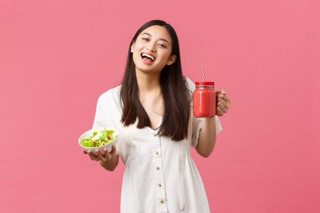 Gesundes Essen, Emotionen und Sommer-Lifestyle-Konzept. Begeistertes und fröhliches, süßes asiatisches Mädchen voller Energie, das leckeren frischen Salat isst und Smoothie trinkt und in die Kamera lächelt glücklicher, rosafarbener Hintergrund.