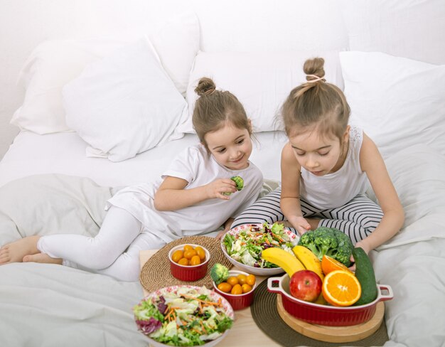 Gesundes Essen zu Hause. Glückliche zwei süße Kinder, die Obst und Gemüse im Schlafzimmer auf dem Bett essen. Gesundes Essen für Kinder und Jugendliche.