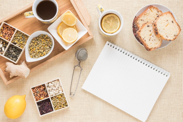 Gesundes Frühstück mit verschiedenen Kräutern; Zitrone; Sieb; Brot; Ingwer und leere Spirale Notizblock