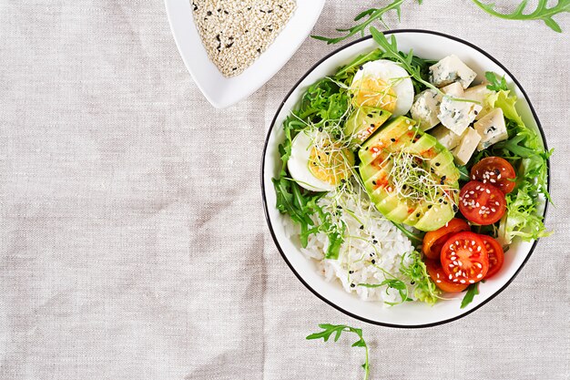 Gesundes grünes vegetarisches Buddha-Schüssel-Mittagessen mit Eiern, Reis, Tomate, Avocado und Blauschimmelkäse auf Tisch.