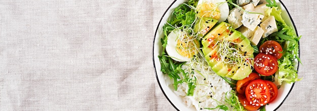 Gesundes grünes vegetarisches Buddha-Schüssel-Mittagessen mit Eiern, Reis, Tomate, Avocado und Blauschimmelkäse auf Tisch.