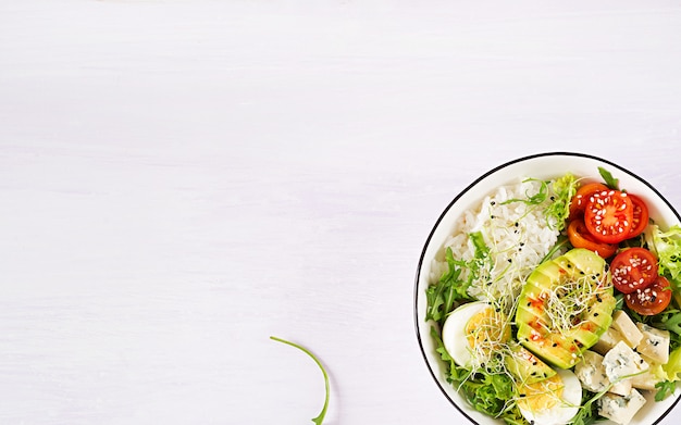 Gesundes grünes vegetarisches Buddha-Schüssel-Mittagessen mit Eiern, Reis, Tomate, Avocado und Blauschimmelkäse auf Tisch.