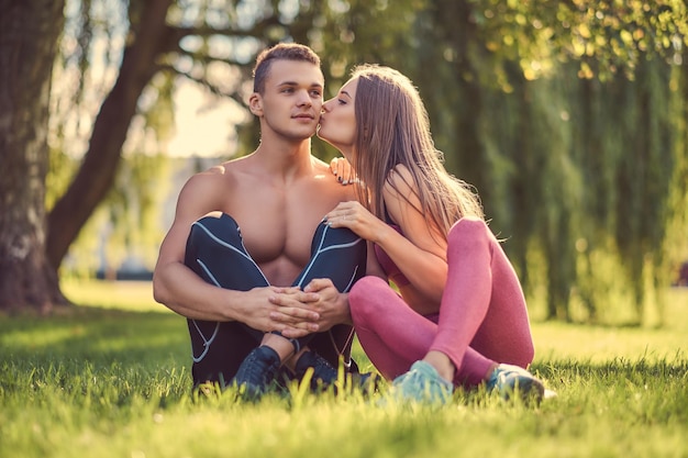 Gesundes Lebensstilkonzept. Glückliches junges Fitnesspaar, das sich beim Sitzen auf einem grünen Gras küsst.