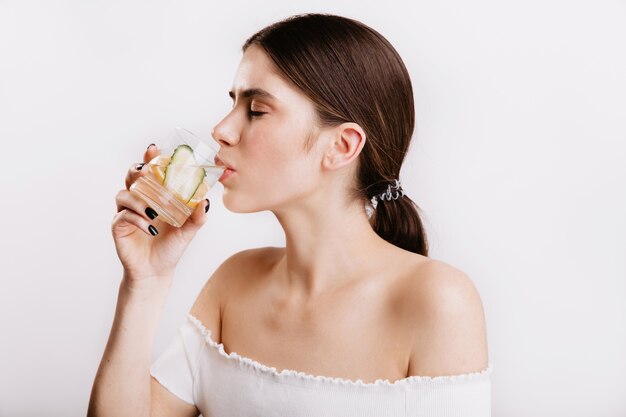 Gesundes Mädchen mit klarer Haut trinkt morgens Wasser mit Zitrone und Gurke. Schuss des schönen Modells ohne Make-up auf weißer Wand.