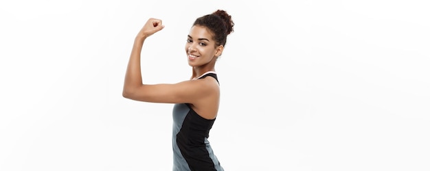 Gesundes und Fitness-Konzept Porträt einer jungen, schönen Afroamerikanerin, die ihre starken Muskeln mit selbstbewusstem, fröhlichem Gesichtsausdruck zeigt, isoliert auf weißem Studiohintergrund