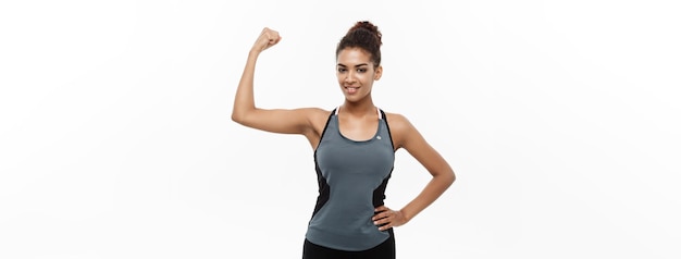 Gesundes und Fitness-Konzept Porträt einer jungen, schönen Afroamerikanerin, die ihre starken Muskeln mit selbstbewusstem, fröhlichem Gesichtsausdruck zeigt, isoliert auf weißem Studiohintergrund