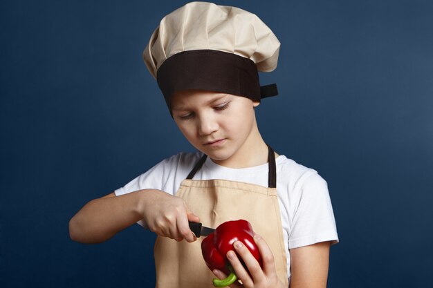 Gesundheit, Ernährung und Ernährung. Bild des ernsthaften konzentrierten kleinen Jungen in der Kochmütze, die an der leeren Wand steht und roten Pfeffer mit Messer schält, während gesundes Abendessen oder Mittagessen mit frischem Gemüse gekocht wird