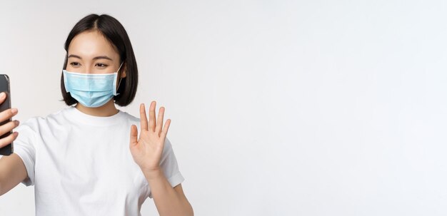 Gesundheits- und covid19-Konzept Moderne asiatische Studentin im Video-Chat mit medizinischer Maske mit Handy, die mit der Hand auf die Smartphone-App winkt, die über weißem Hintergrund steht