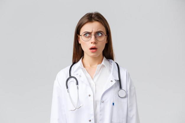 Gesundheitspersonal, Medizin, Versicherung und Covid-19-Pandemiekonzept. Schockierte und verwirrte junge Ärztin in weißen Kitteln und Brille, Stethoskop, starrte mit besorgtem nervösem Gesicht nach links