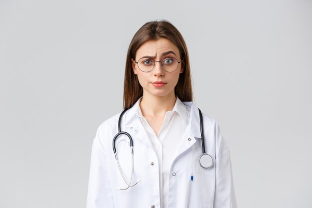 Gesundheitspersonal, Medizin, Versicherung und Covid-19-Pandemiekonzept. Skeptische und verwirrte Ärztin in weißen Kitteln, medizinischem Anzug und Brille, Augenbrauen urteilend hochziehen, unzufrieden grinsen
