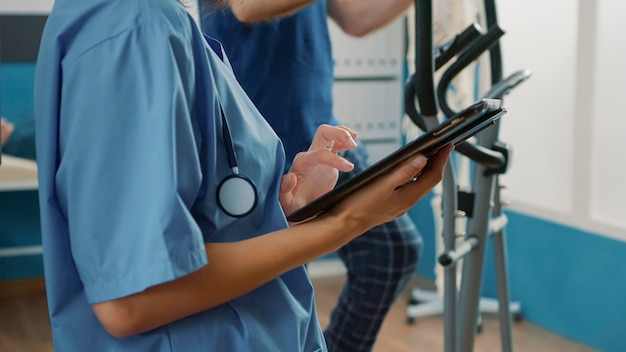 Gesundheitsspezialist in Uniform mit digitalem Tablet, um älteren Patienten mit körperlichen Verletzungen und mechanischen Störungen zu helfen. Krankenschwester-Haltevorrichtung bei Muskelrehabilitationsverfahren. Nahansicht.