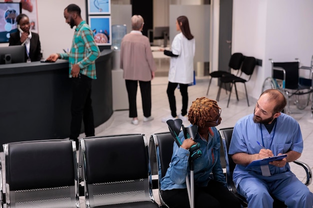 Gesundheitsspezialistin berät Frau mit Krücken in der Empfangshalle des Krankenhauses und spricht über Diagnose und Medizin im Wartezimmerbereich. Diverse Leute diskutieren beim Kontrolltermin.