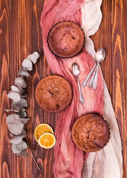 Getrocknete Eukalyptuspopulusblätter und Zitrusfruchtscheiben mit drei gebackenen Kuchen und Löffeln auf Leinen kleidet über dem Holztisch