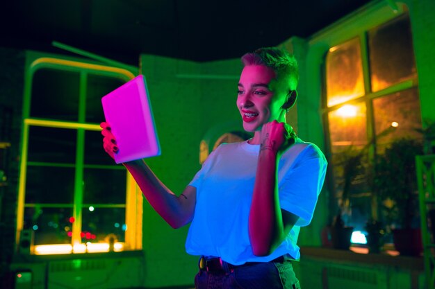 Gewinnen. Filmporträt der stilvollen Frau im neonbeleuchteten Innenraum. Getönt wie Kinoeffekte, leuchtende Neonfarben. Kaukasisches Modell mit Tablette in bunten Lichtern drinnen. Jugendkultur.