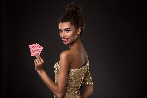 Gewinnende Frau - Junge Frau in einem eleganten goldenen Kleid mit zwei Karten, einer Kombination aus Assen und Pokerkarten. Studioaufnahme auf schwarzem Hintergrund. Eine junge Frau steht mit dem Rücken