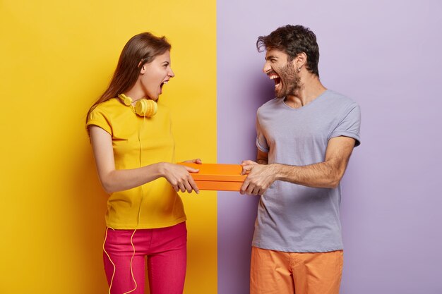 Gierige Frauen und Männer können sich keine Schachtel teilen, beide halten ein orangefarbenes Päckchen, schreien sich gegenseitig an, haben genervte Gesichtsausdrücke, tragen farbenfrohe Kleidung und stehen vor zweifarbigem Hintergrund.