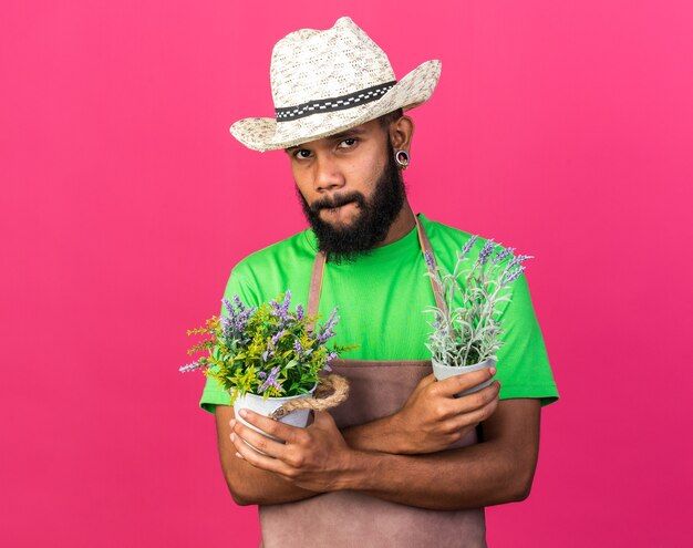 Gieriger junger Gärtner afroamerikanischer Typ mit Gartenhut, der Blumen im Blumentopf hält und kreuzt