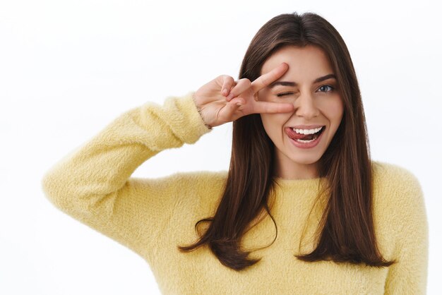 Glänzen Sie hell Fröhliche, schöne weibliche Frau, die kawaii Friedensgeste auf dem Auge zeigt und ein lächelndes, freches Kameramädchen ausdrückt, das glückliche und positive Emotionen ausdrückt, hat keine Sorgen, steht entspannt und optimistisch