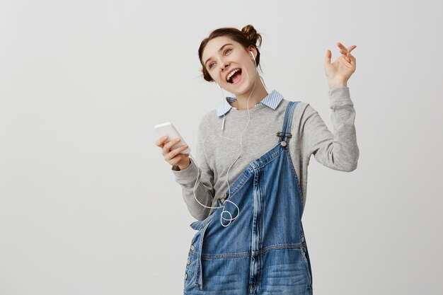 Glänzendes erwachsenes Mädchen mit braunen Haaren, die wie Stern handeln, der neuen entzückenden Titel vom Smartphone hört. Fröhliche Frau singt ekstatisch, während sie Freizeit verbringt. Zeitvertreibskonzept