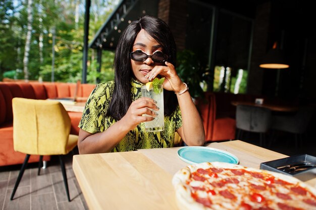 Glamour-Afroamerikanerin, die im Restaurant Pizza isst und Limonade trinkt