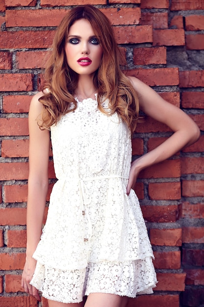 Glamour-Schönheitsporträt des schönen sinnlichen kaukasischen jungen Frauenmodells mit Abendmake-up im weißen Sommerkleid, das auf der Straße nahe Ziegelmauer aufwirft