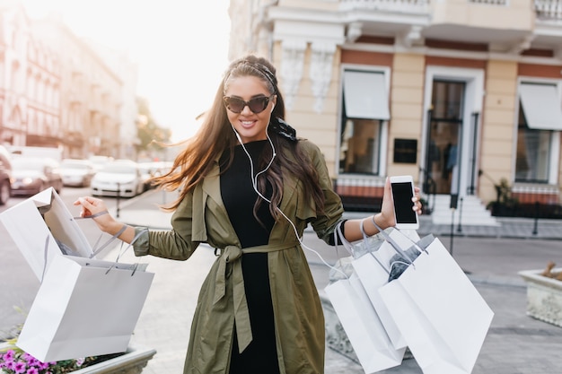 Glamouröse dunkelhaarige Dame mit Smartphone, die nach dem morgendlichen Einkauf die Straße entlang geht