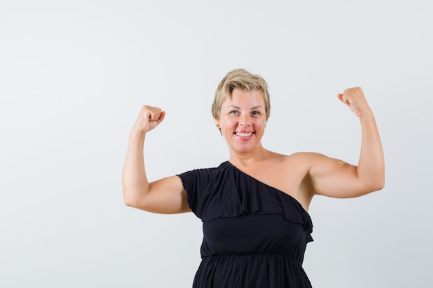 Glamouröse Frau, die ihre Armkraft in der schwarzen Bluse zeigt und fröhlich aussieht. Vorderansicht.