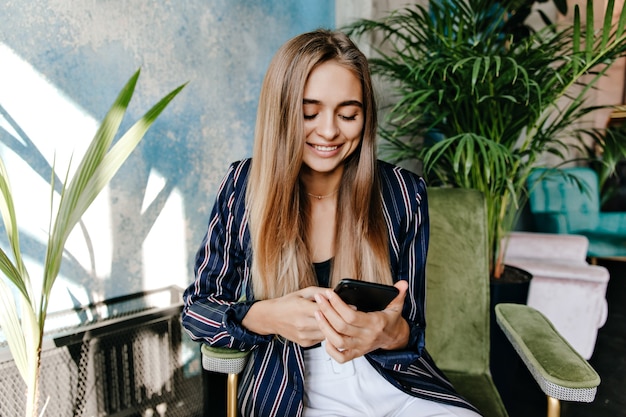 Glamouröse Frau, die Telefonbildschirm beim Sitzen im Büro betrachtet. Innenaufnahme des erstaunlichen hübschen Mädchens, das im Sessel ruht.