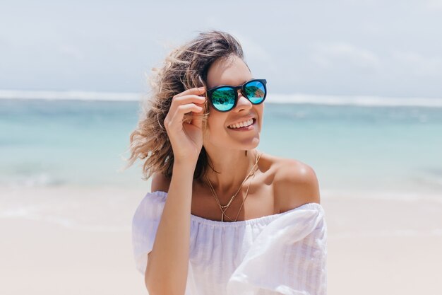 Glamouröse Frau im weißen Kleid, die Sommer im Resort genießt. Porträt der atemberaubenden gebräunten Dame in der Sonnenbrille, die nahe Meer steht.