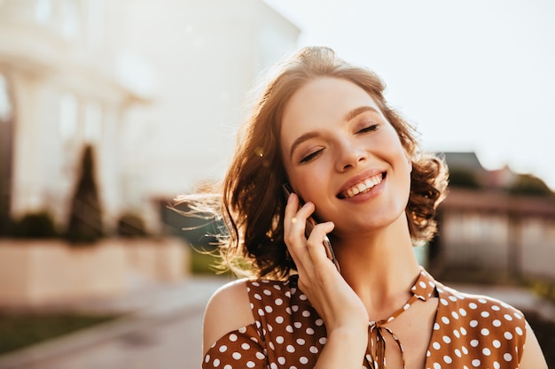 Glamouröse junge Frau, die mit geschlossenen Augen am Telefon spricht. Außenaufnahme des hübschen kaukasischen Mädchens mit den kurzen braunen Haaren.