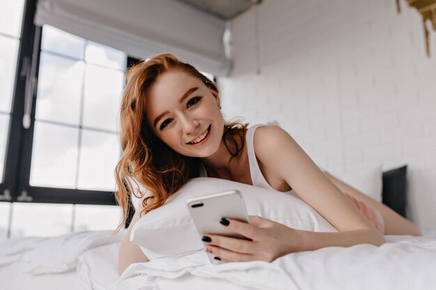 Glamouröse junge Frau mit schwarzer Maniküre, die im Bett liegt. Lächelndes niedliches Ingwermädchen, das im Schlafzimmer mit Smartphone aufwirft.
