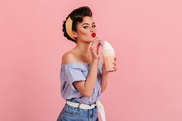 Glamouröse kaukasische Frau, die Milchshake trinkt. Studioaufnahme des modischen brünetten Mädchens, das mit Cocktail auf rosa Hintergrund aufwirft.