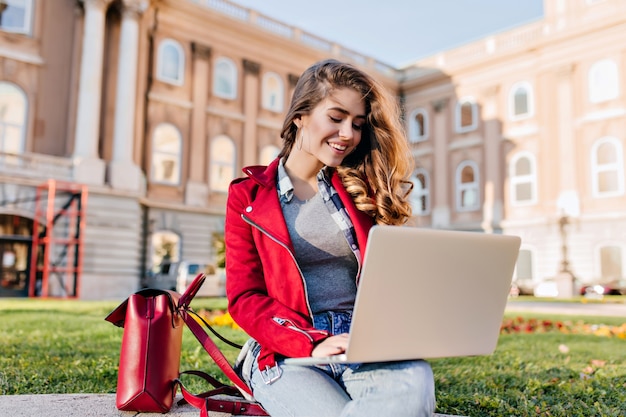 Glamouröse Studentin in roter Jacke, die im Hof vor dem College mit Computer sitzt