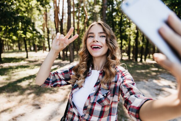 Glamouröses lockiges Mädchen, das mit Lächeln im Wald aufwirft. Charmantes weibliches Modell mit Smartphone für Selfie auf Natur.