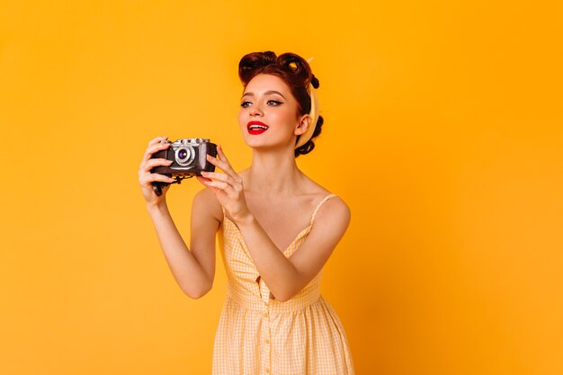 Glamouröses Pinup-Girl beim Fotografieren. Inspirierte Ingwerfrau mit der Kamera, die auf gelbem Raum steht.