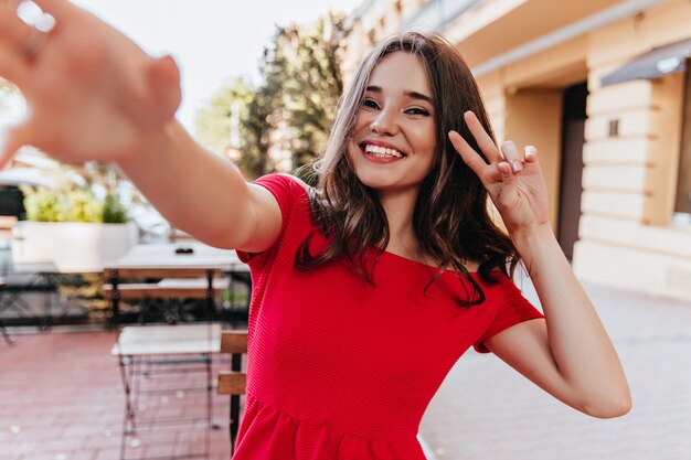 Glamouröses weißes Mädchen, das im Straßencafé herumalbert. Wunderschönes brünettes weibliches Modell, das Selfie am Sommertag macht.