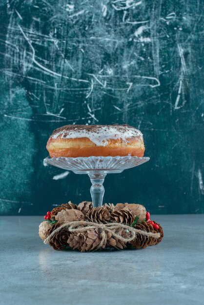 Glasierter Donut auf einem Glassockel inmitten eines Kiefernkranzes auf Marmor.